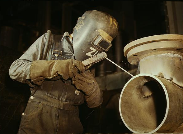 Historical photograph of a welder at work in protective helmet, gloves, and overalls.